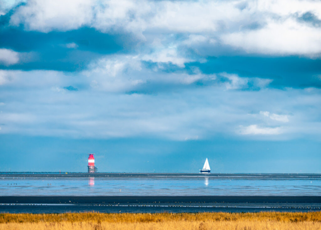 Foto eines Segelschiffes und eines Leuchtturmes im blauen Meer vor Wolkenverhangenem Himmel