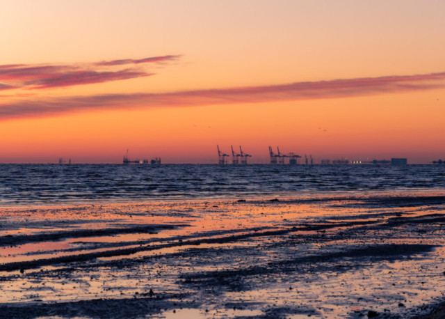 Foto eines Sonnenaufgangs am Meer. Am Horizont sieht man Schiffskräne und andere Hafengebäude