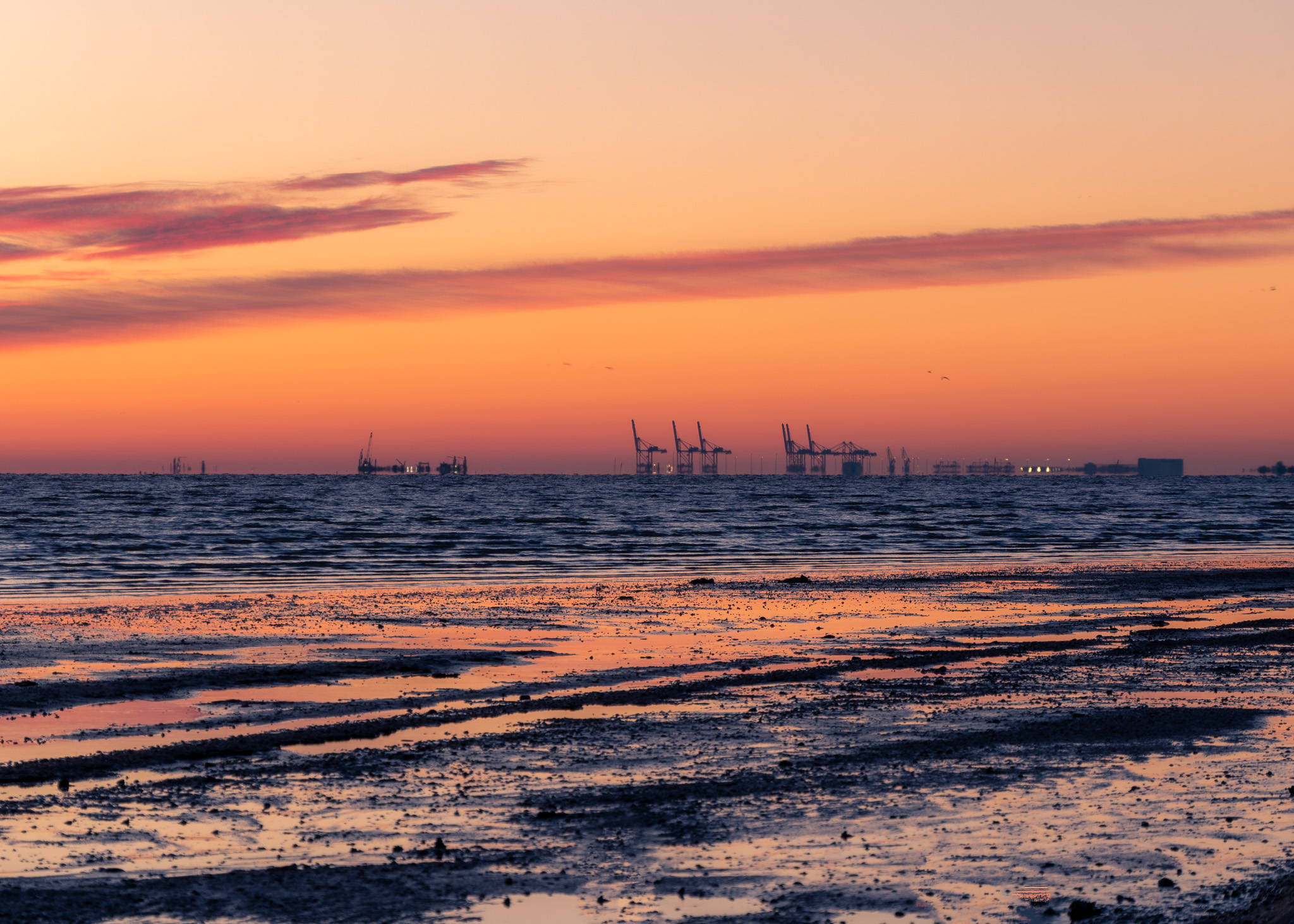 Foto eines Sonnenaufgangs am Meer. Am Horizont sieht man Schiffskräne und andere Hafengebäude
